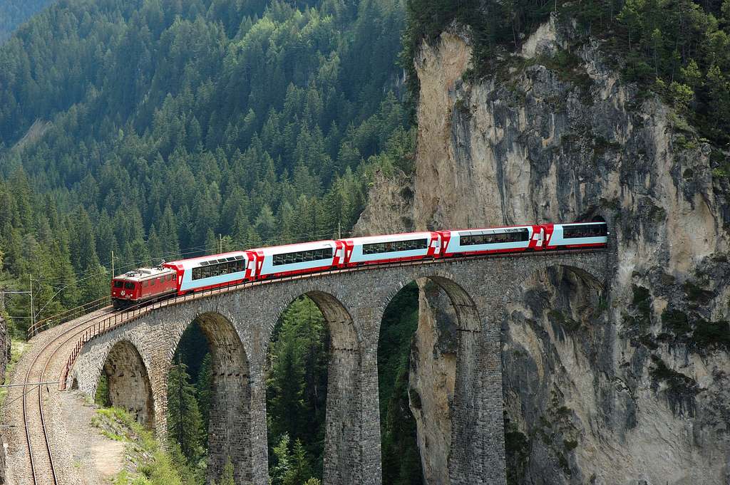 Fotka zobrazuje vlak, ktorý prechádza práve po moste a okolo neho je hornatá krajina s veľkým množstvom stromov. Vlak je bielo-červený.